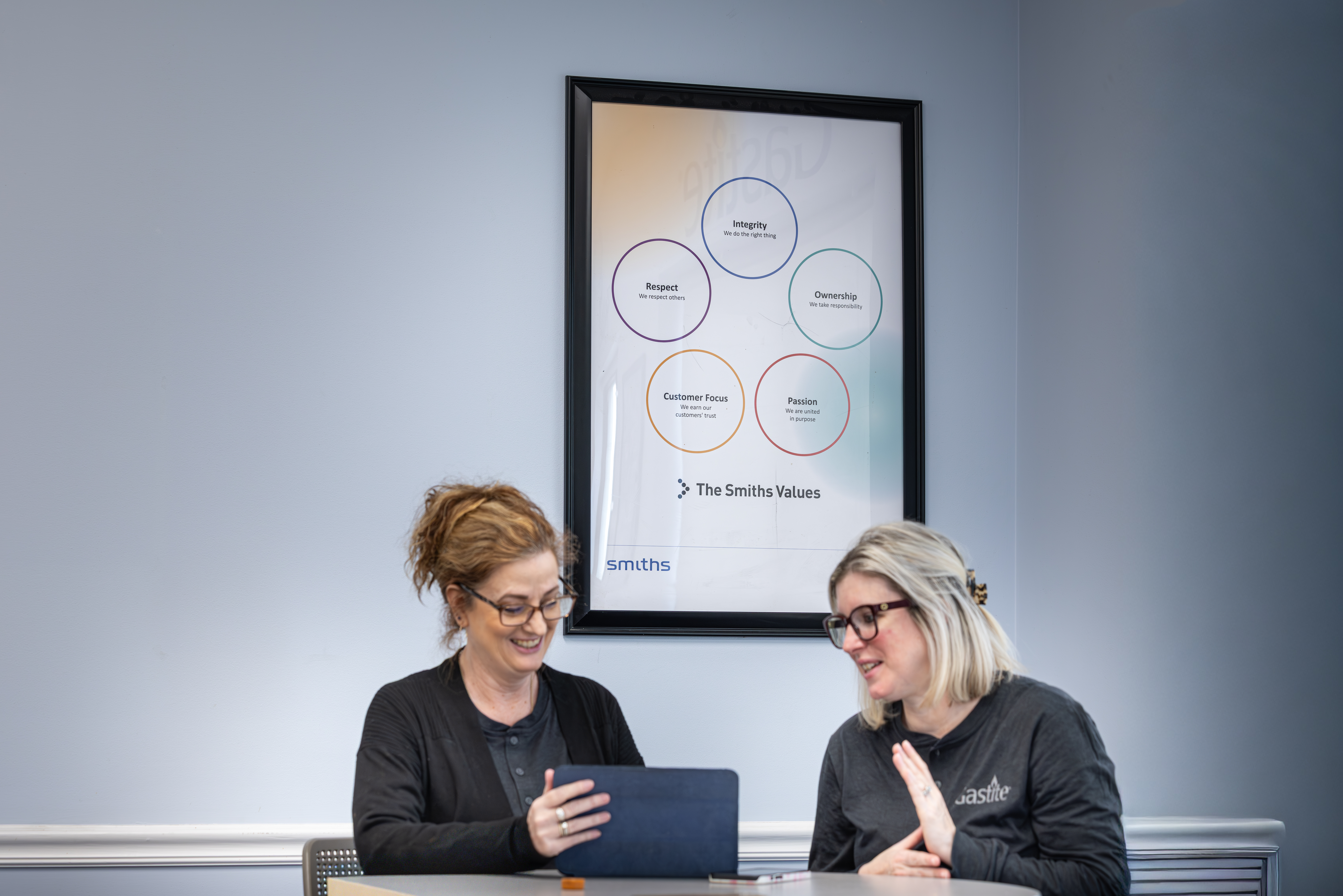 Two Women In Gastite Uniforms Smiling While Looking At A Tablet Smiths Value Poster In Background