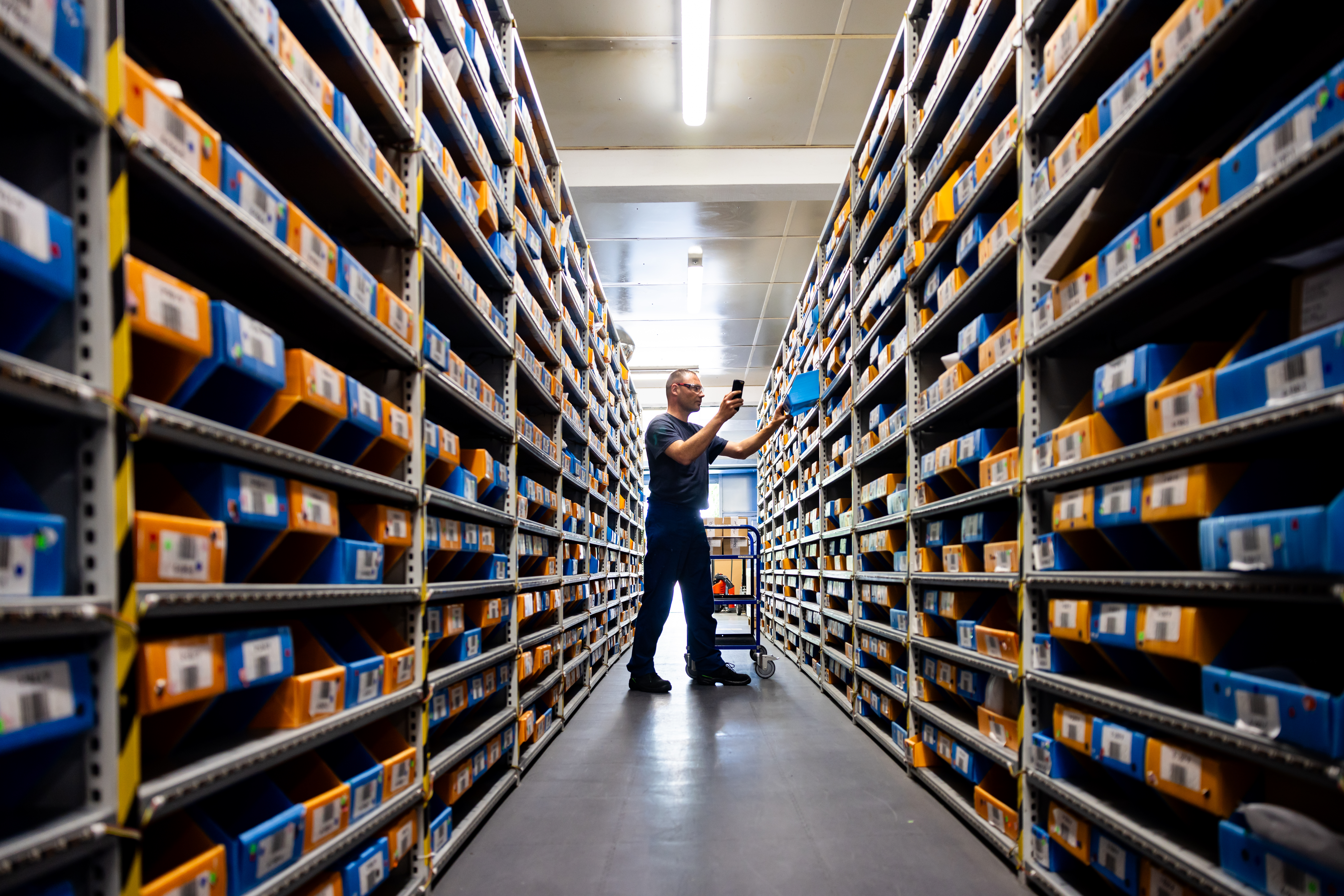 Worker In Warehouse Between Rows Of Shelves